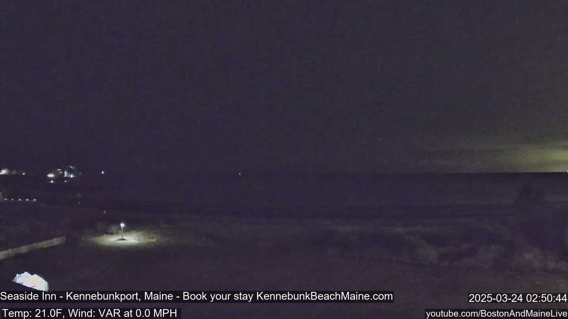 A nighttime view of a dark ocean shore with a single lit lamppost in the foreground under a clear, dark sky.
