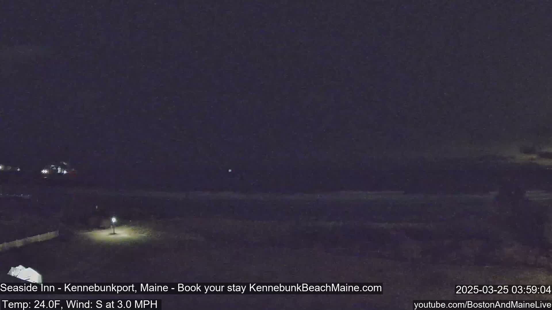 A nighttime view of a dark beach and shoreline under a clear, dark sky with a single lamp lit in the foreground.