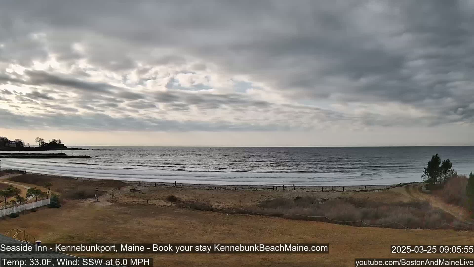 An overcast, breezy day at a beach shows a calm ocean with small waves rolling onto a sandy shore, with some vegetation and a small rocky point in the distance.