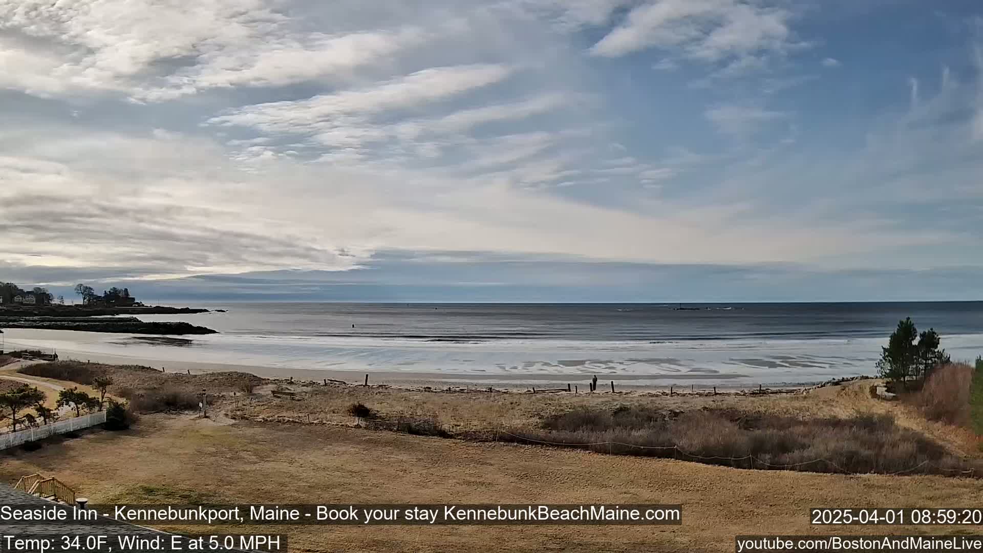 A mostly cloudy day reveals a beach with patches of ice, calm waves lapping the shore, and a few people walking along the sand.