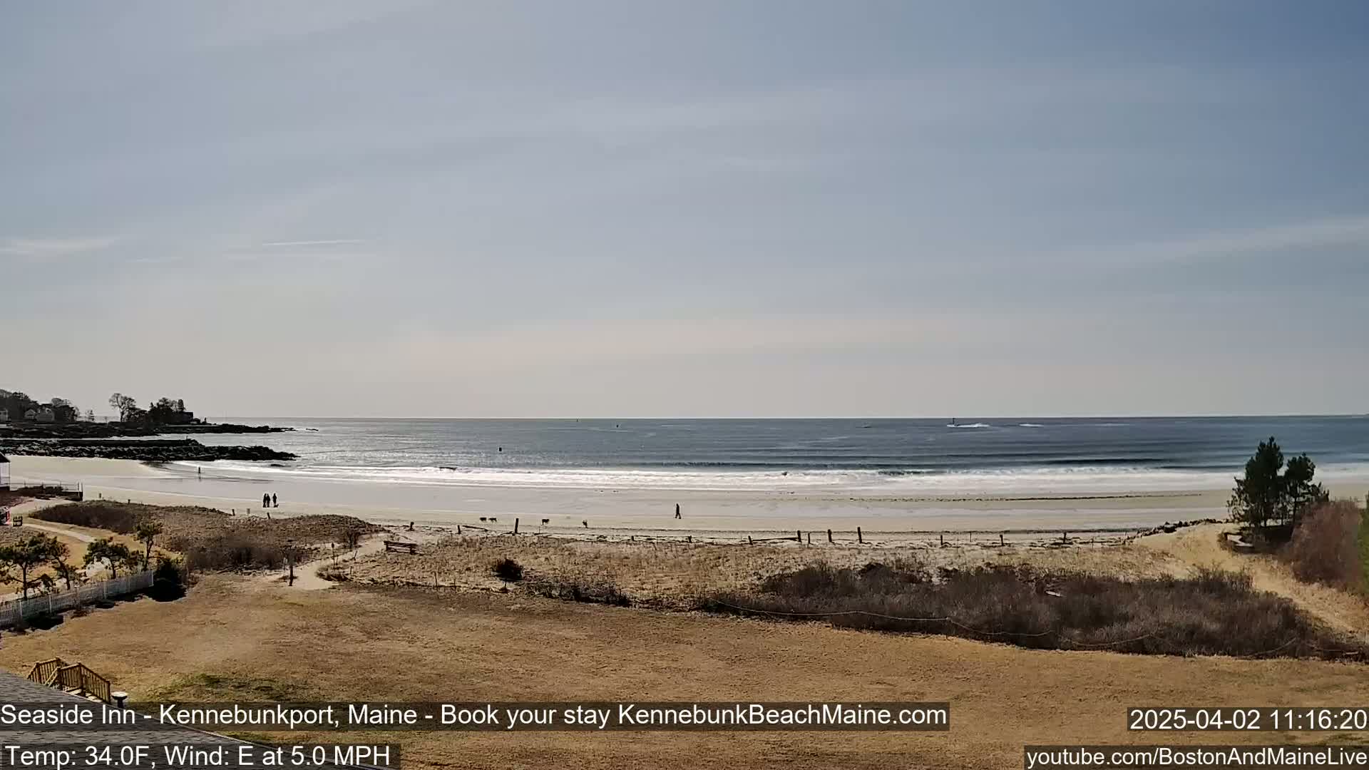 A sandy beach with gentle waves and a few people walking, under a clear, sunny sky.