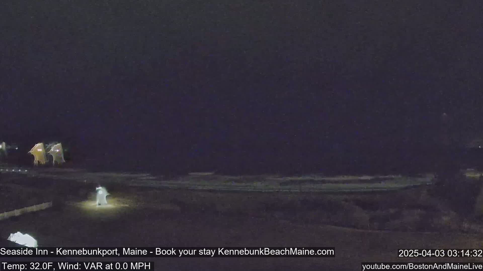 A nighttime view of a dark beach with dimly lit buildings in the distance under a mostly clear sky.