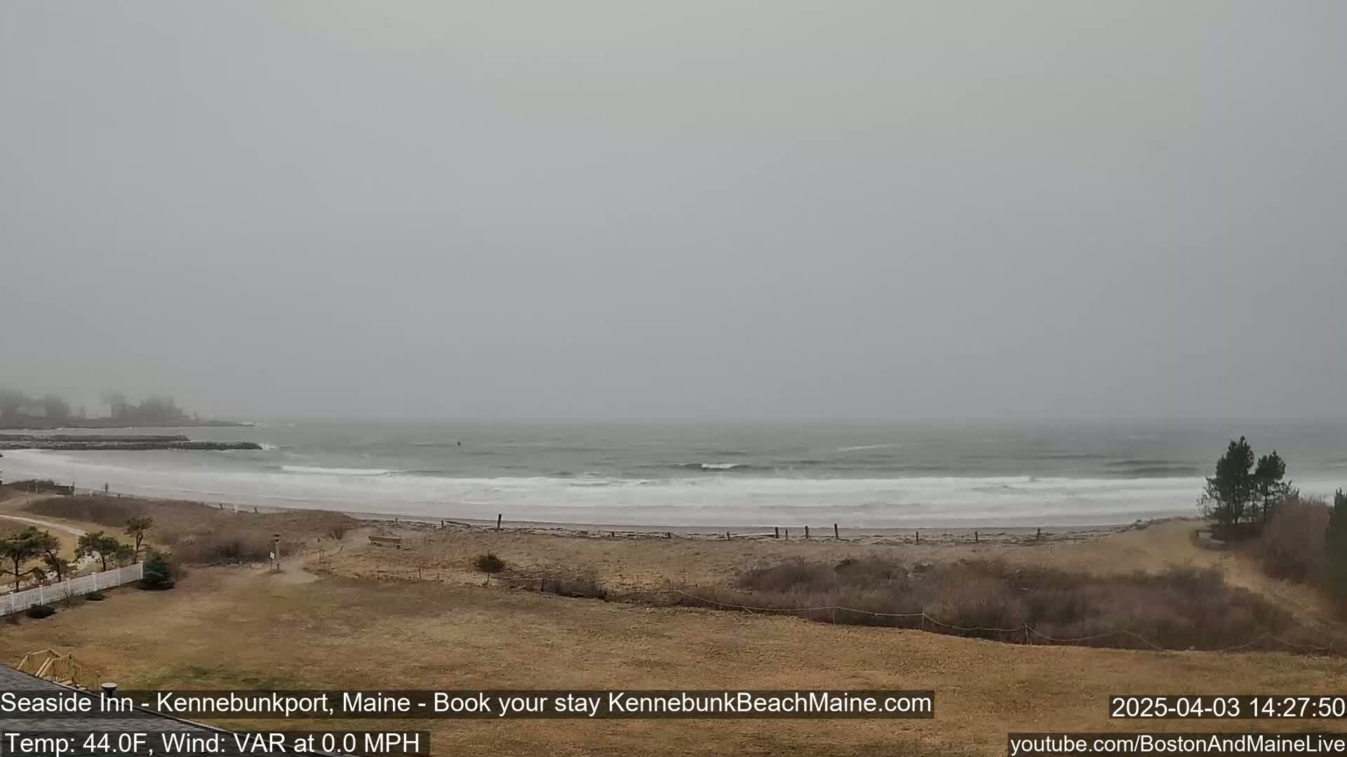 A hazy, overcast day shows a sandy beach with small waves lapping the shore, some vegetation, and a glimpse of buildings in the distance.