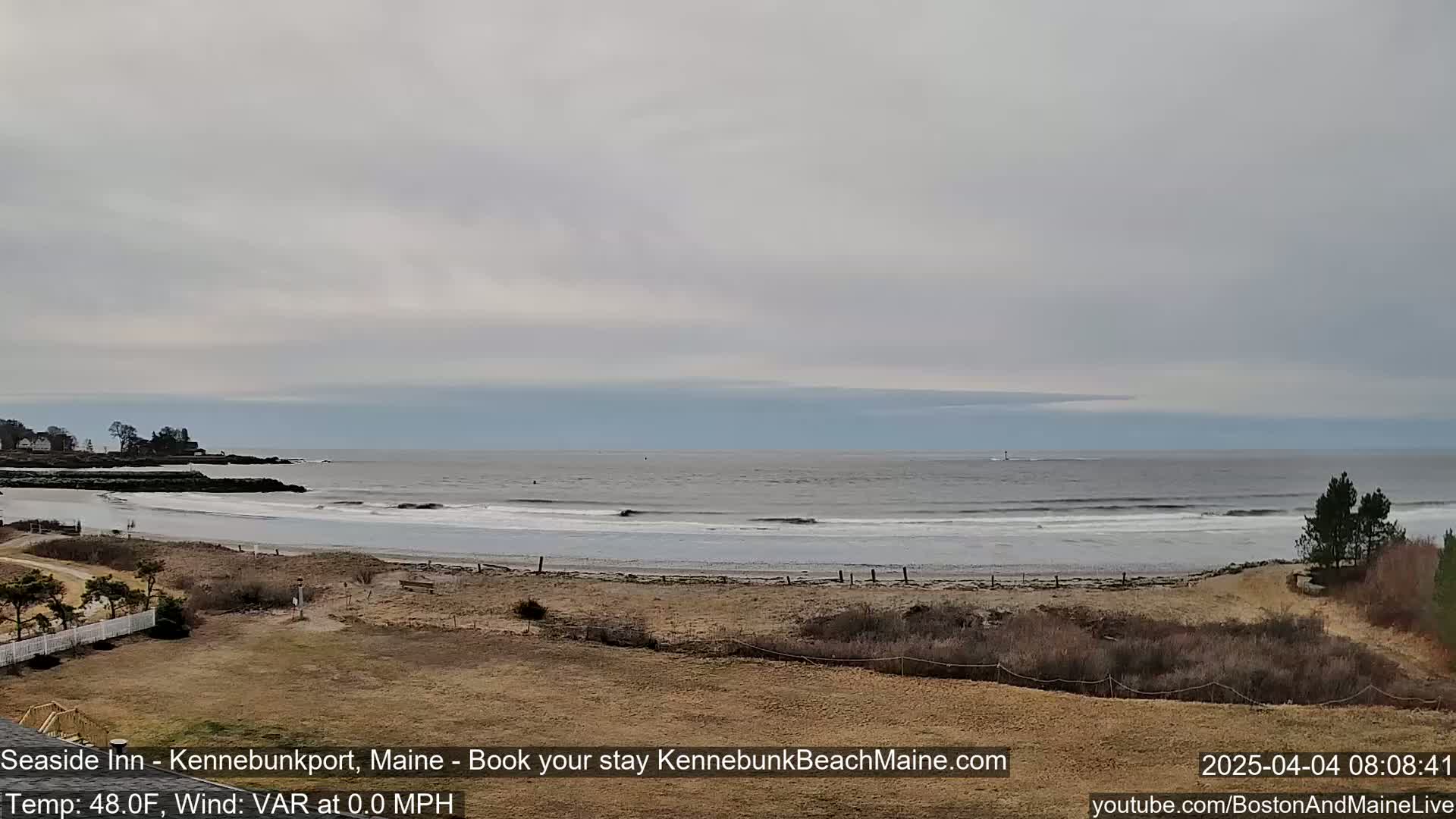 A mostly cloudy day reveals a beach with small waves gently lapping the shore, backed by sparse vegetation and a distant, quiet town.