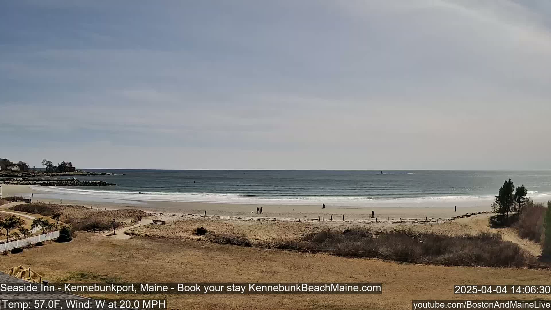 A sandy beach with a few people on it stretches along a calm ocean under a mostly clear sky, with a moderate wind blowing from the west.