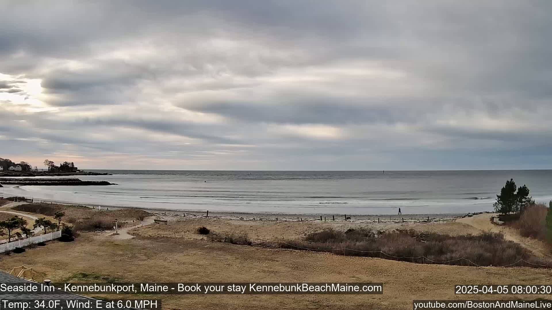 A mostly cloudy day reveals a long, sandy beach with gentle waves lapping the shore, backed by sparse, low-lying vegetation and a few distant houses.