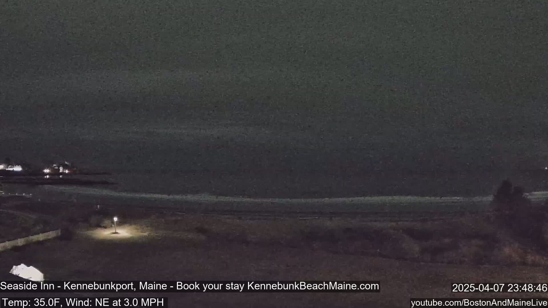 A dark nighttime view of a beach and ocean under a cloudy sky, with a single lit lamppost visible in the foreground.