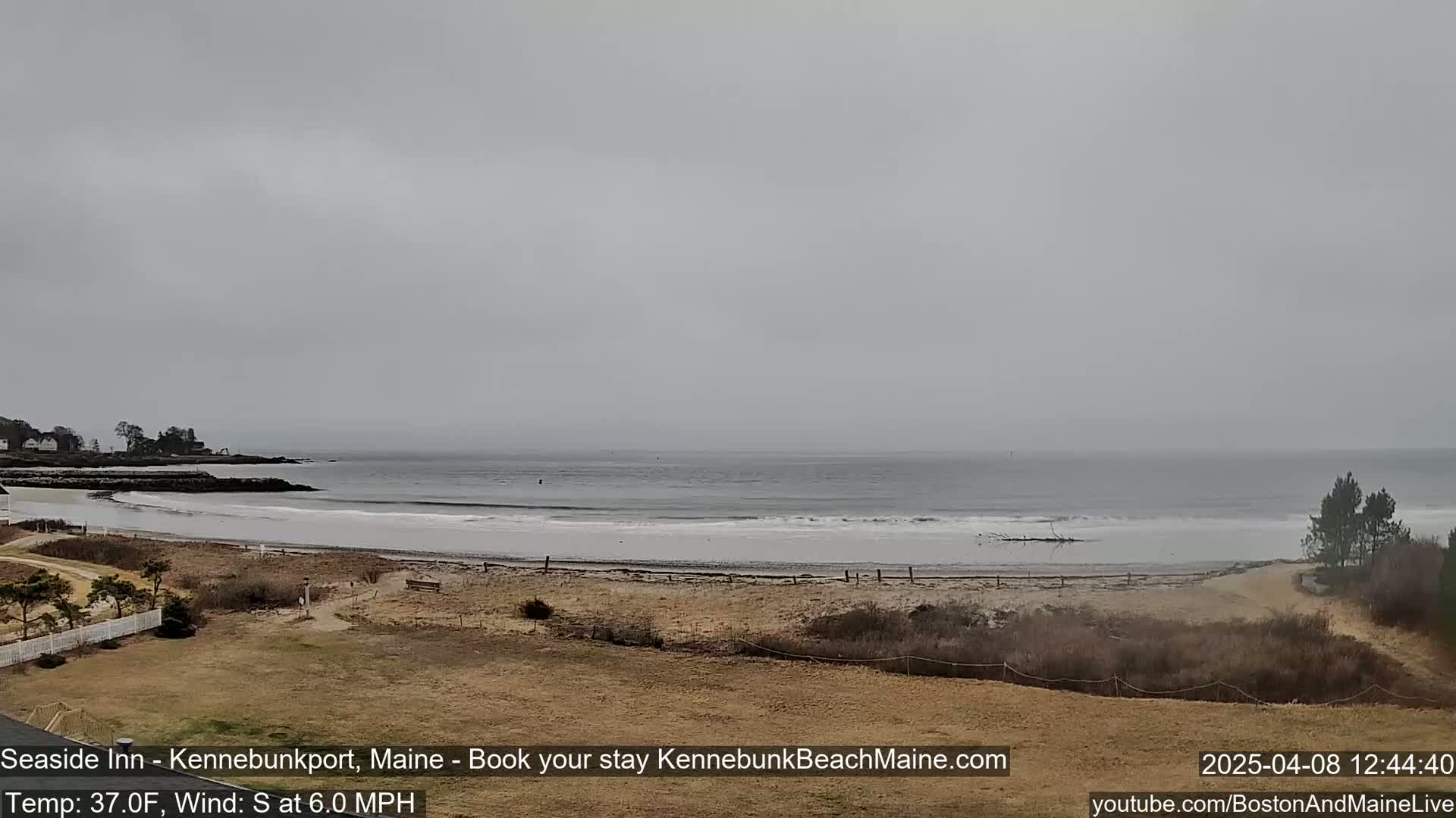 A mostly overcast day reveals a sandy beach meeting a calm ocean, backed by sparse, brown vegetation and a few distant houses.