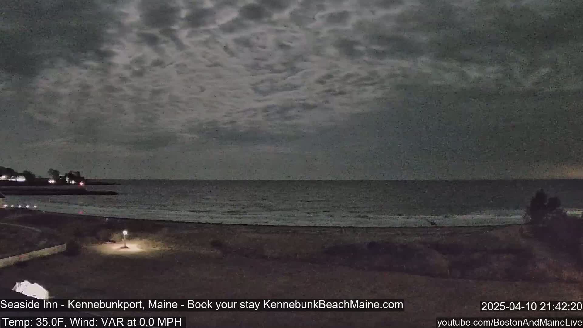 A dark, overcast night scene shows a calm ocean meeting a sandy beach, with a few lights visible on the shore.
