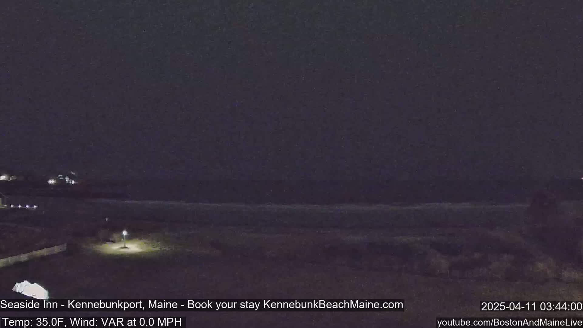 A nighttime view of a dark beach with a single lit lamppost, calm ocean, and a distant dark shoreline under a clear, dark sky.
