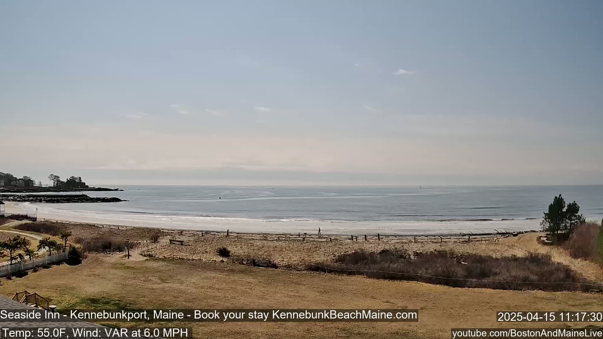 A sandy beach stretches along a calm ocean under a mostly clear sky, with a gentle wind.