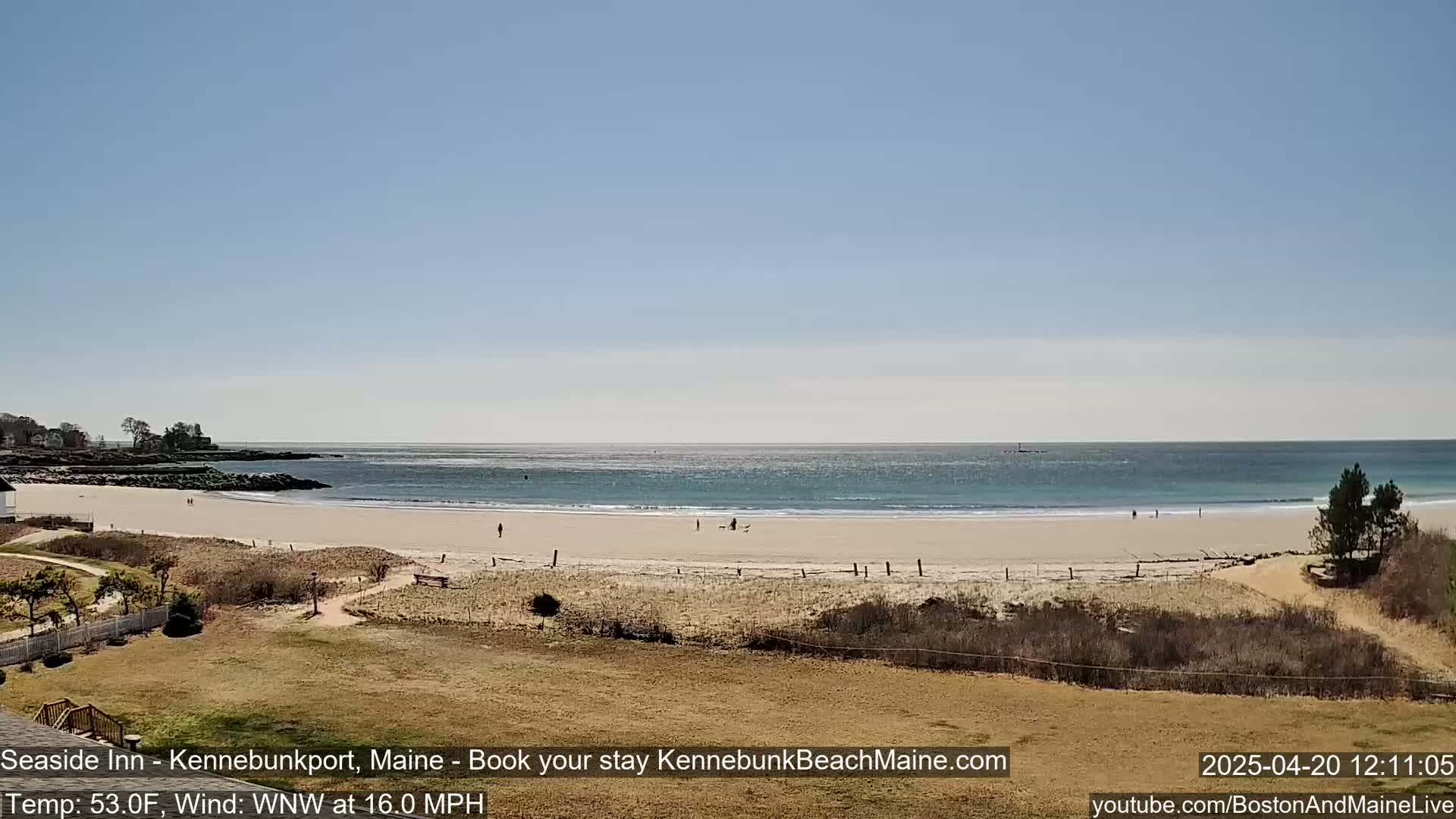 A sandy beach with a few people on it stretches along a calm ocean under a clear blue sky.