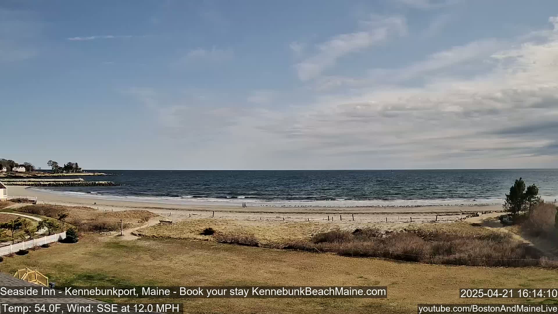 A sandy beach with a calm ocean under a partly cloudy sky is seen from a grassy hill.