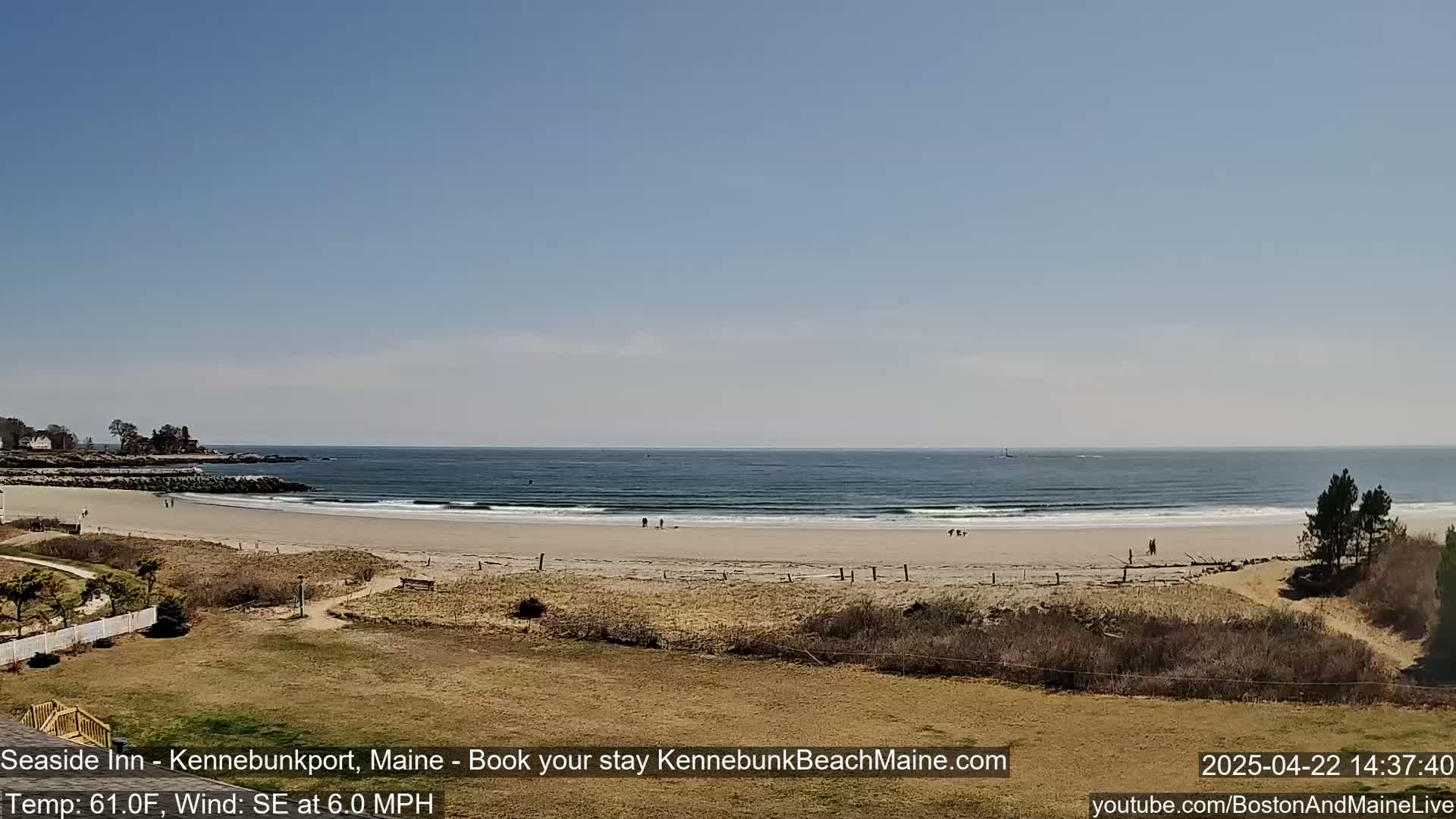 A sandy beach with a calm ocean under a clear blue sky is viewed from a slight elevation, showing small groups of people scattered along the shore.