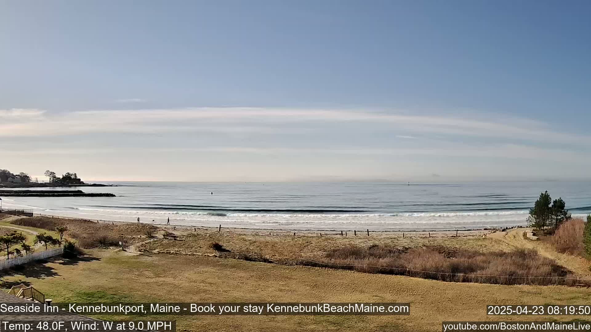 A mostly sunny day at a beach shows calm waves lapping a sandy shore, with a few people walking along the beach and some buildings visible in the distance.
