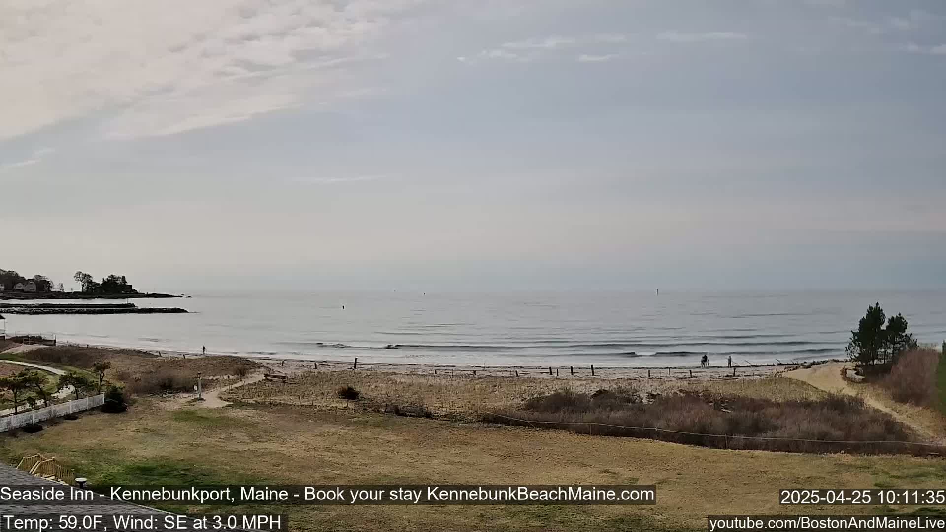 A sandy beach with small waves and a few people stretches along a calm ocean under a mostly cloudy sky.