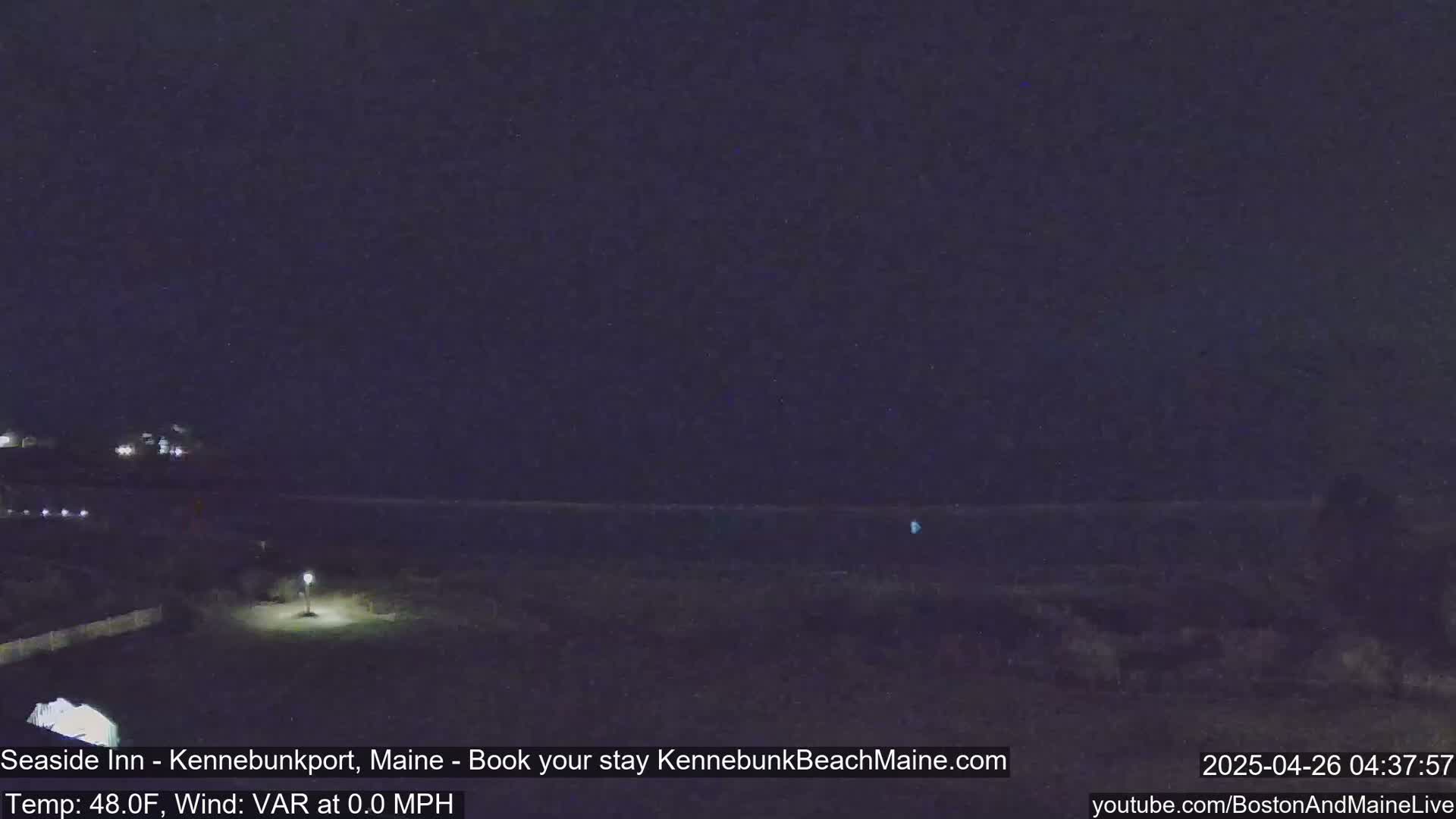 A nighttime view of a dark coastline under a clear, dark sky shows a calm ocean and a dimly lit beachfront area.