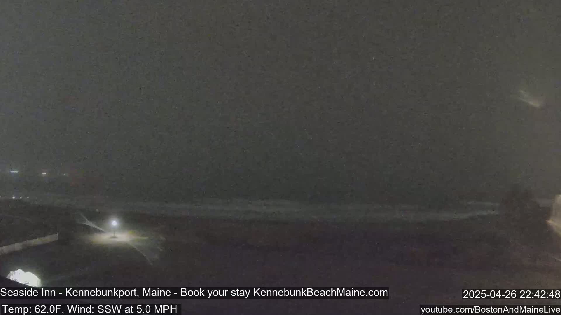 A nighttime, dark view of a beach with a light post and some small waves under a mostly cloudy sky with a slight south-southwest wind at 5 mph.
