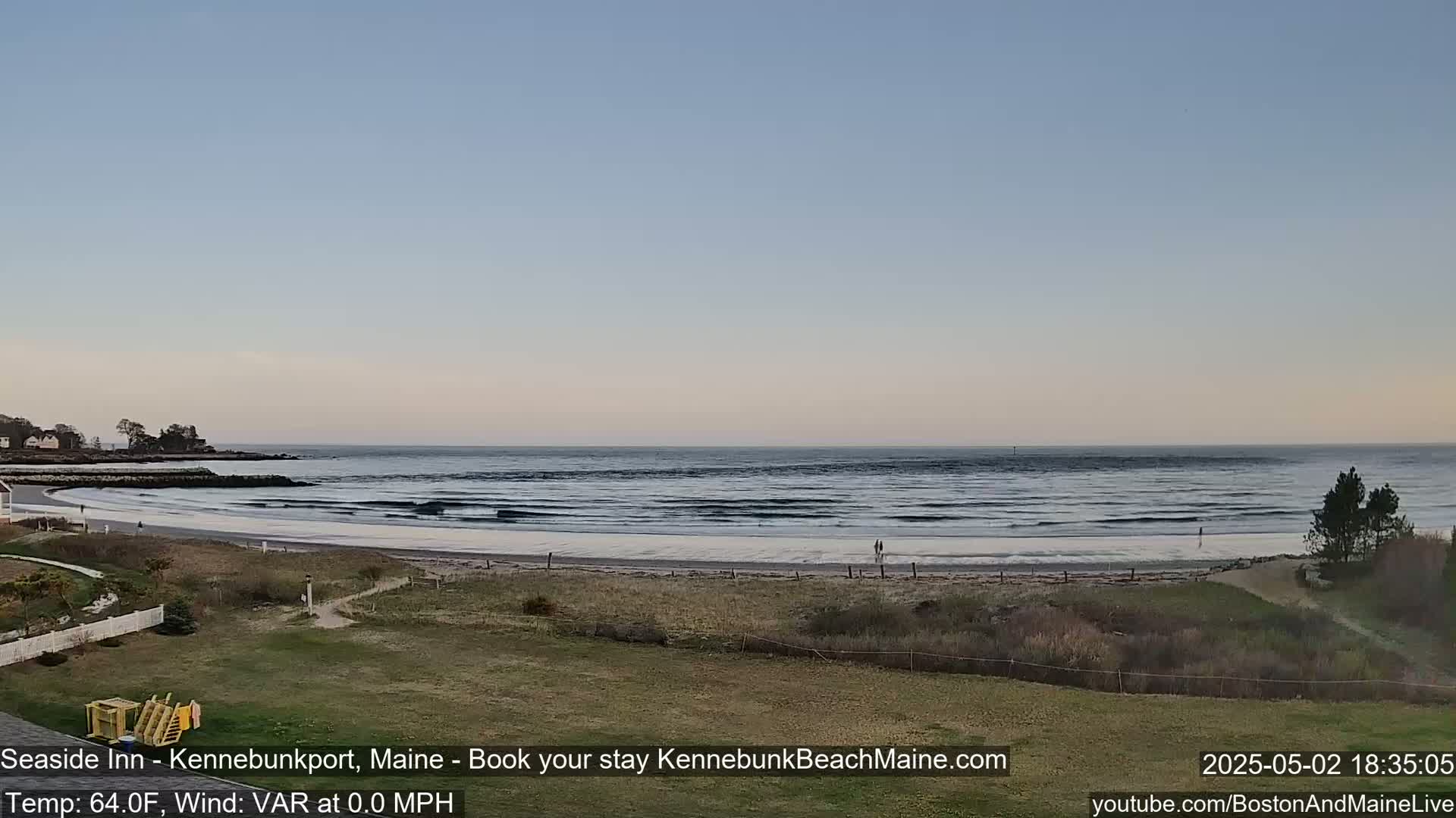 A calm ocean with small waves laps a sandy beach, under a clear, light-blue sky.