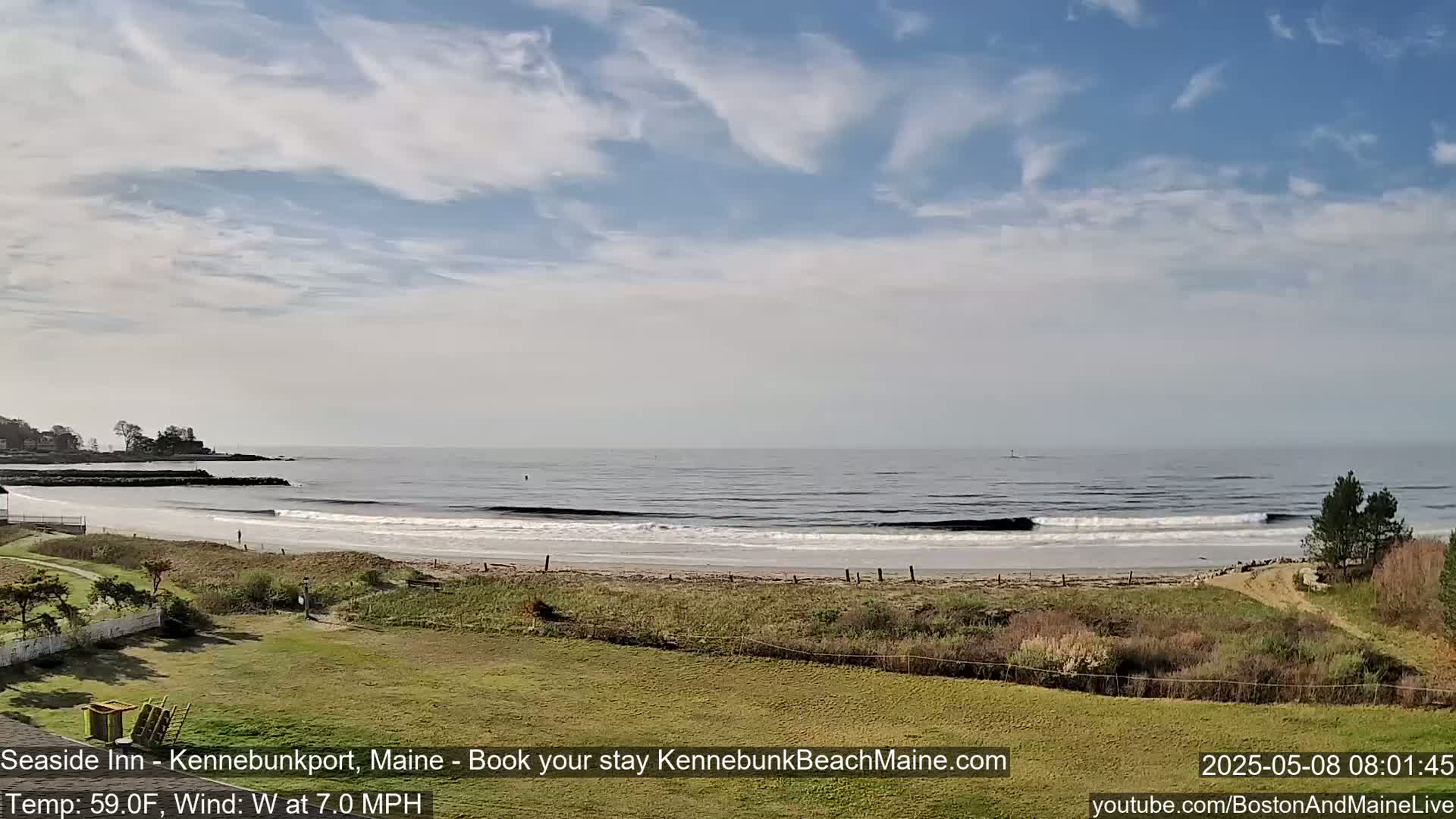 A partly cloudy day shows a sandy beach with small waves lapping the shore, backed by grassy dunes and a distant coastline.