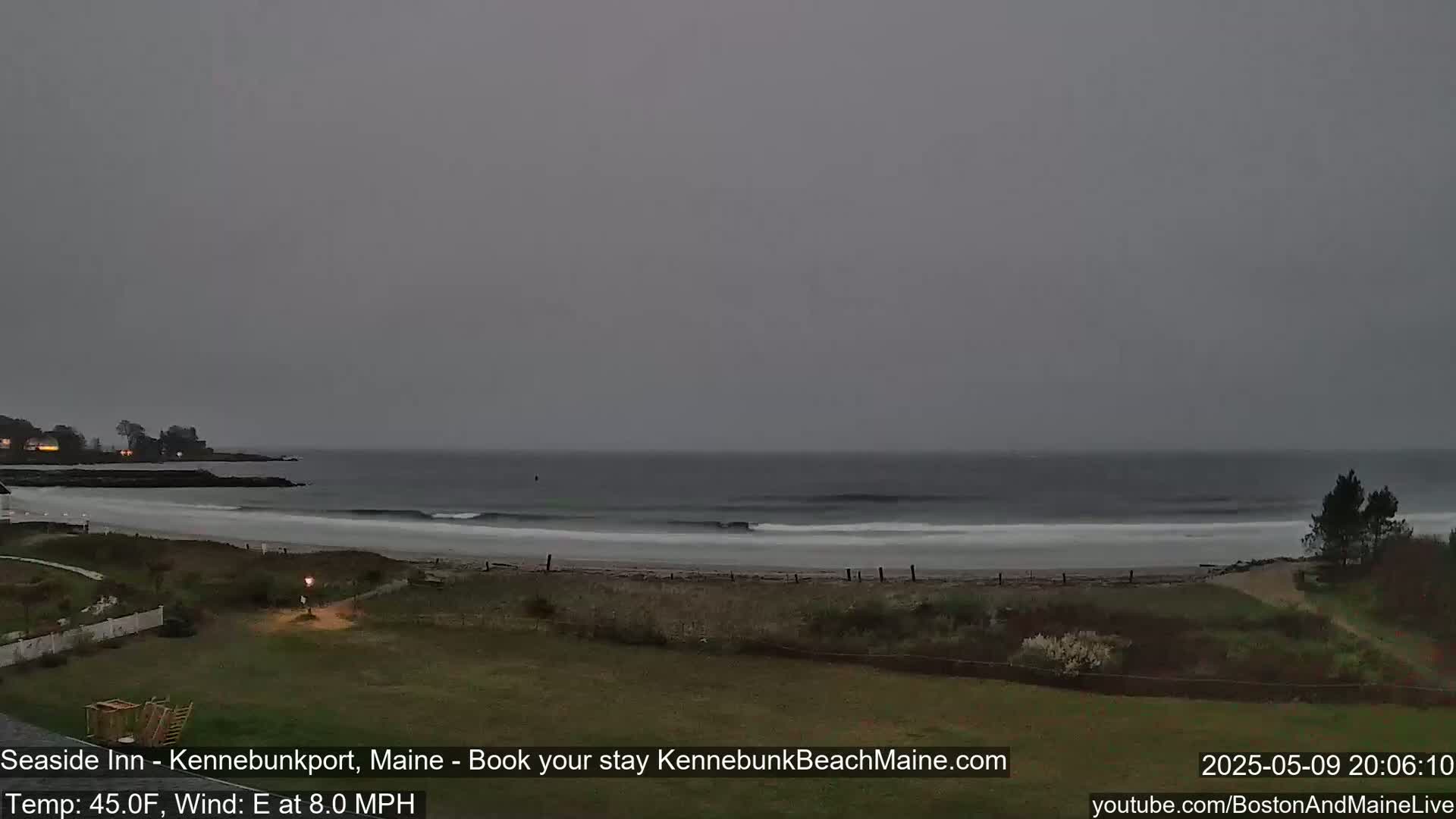 A gray, overcast day reveals a beach scene with gentle waves rolling onto the shore, a grassy area in the foreground, and a distant coastline.
