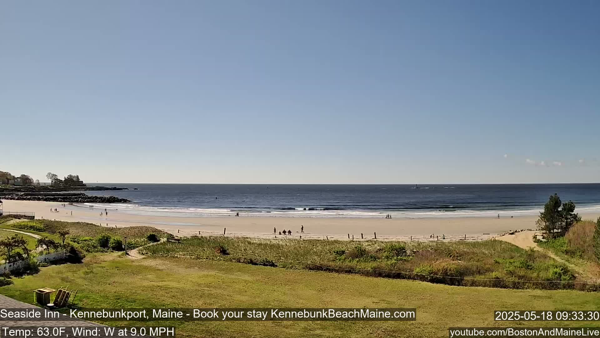A sandy beach with a few people on it stretches along a calm ocean under a clear blue sky.