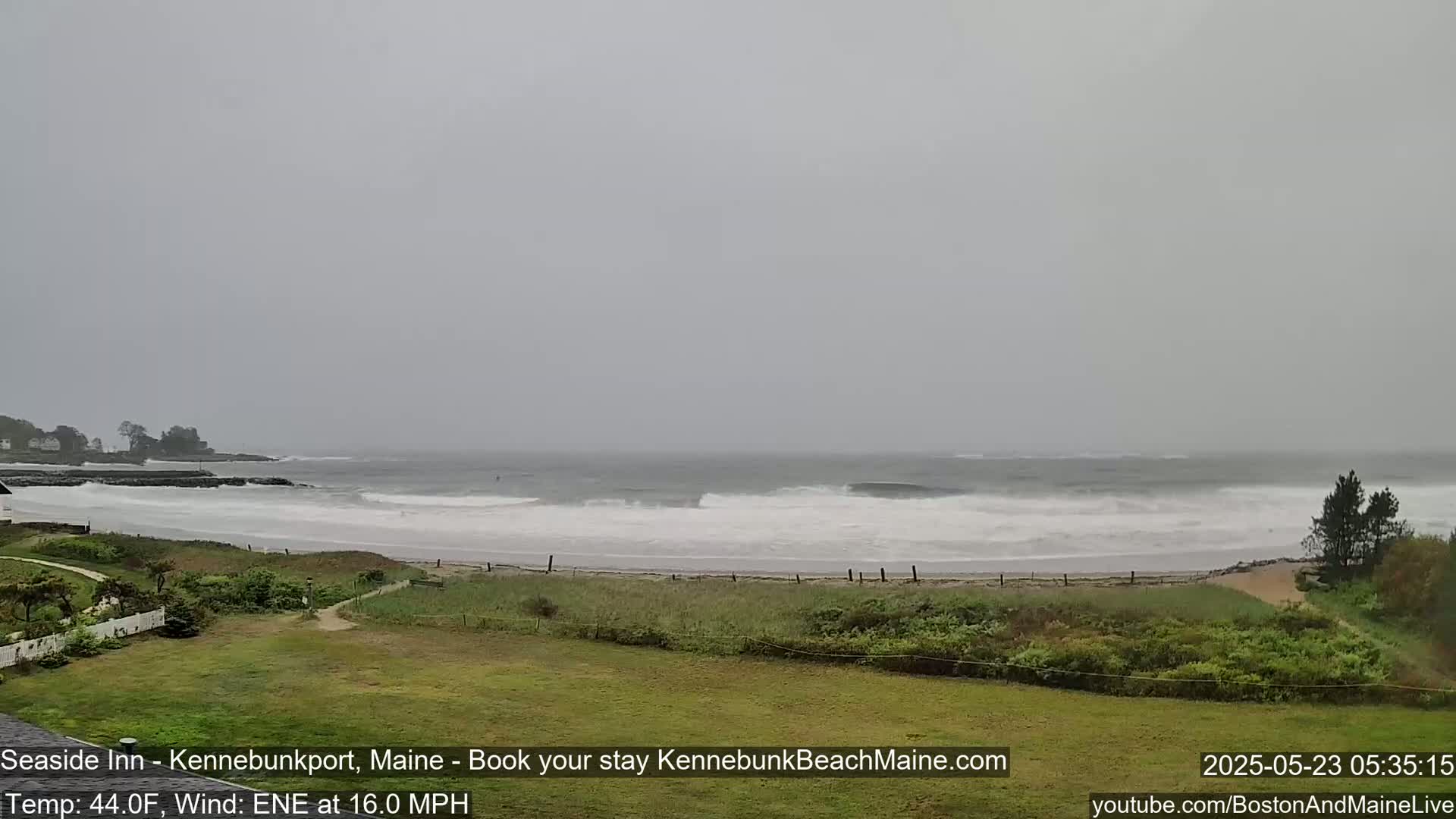 A gray, overcast day shows a beach with moderate waves crashing on the shore, a grassy area in the foreground, and some trees and buildings in the distance.