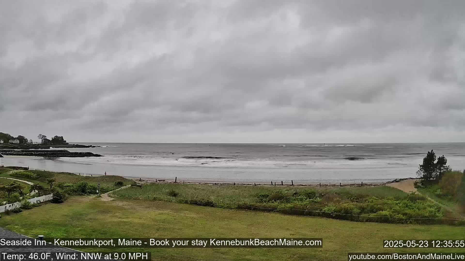 A gray, overcast day shows a view of a beach with calm waves, a grassy area, and some distant buildings.