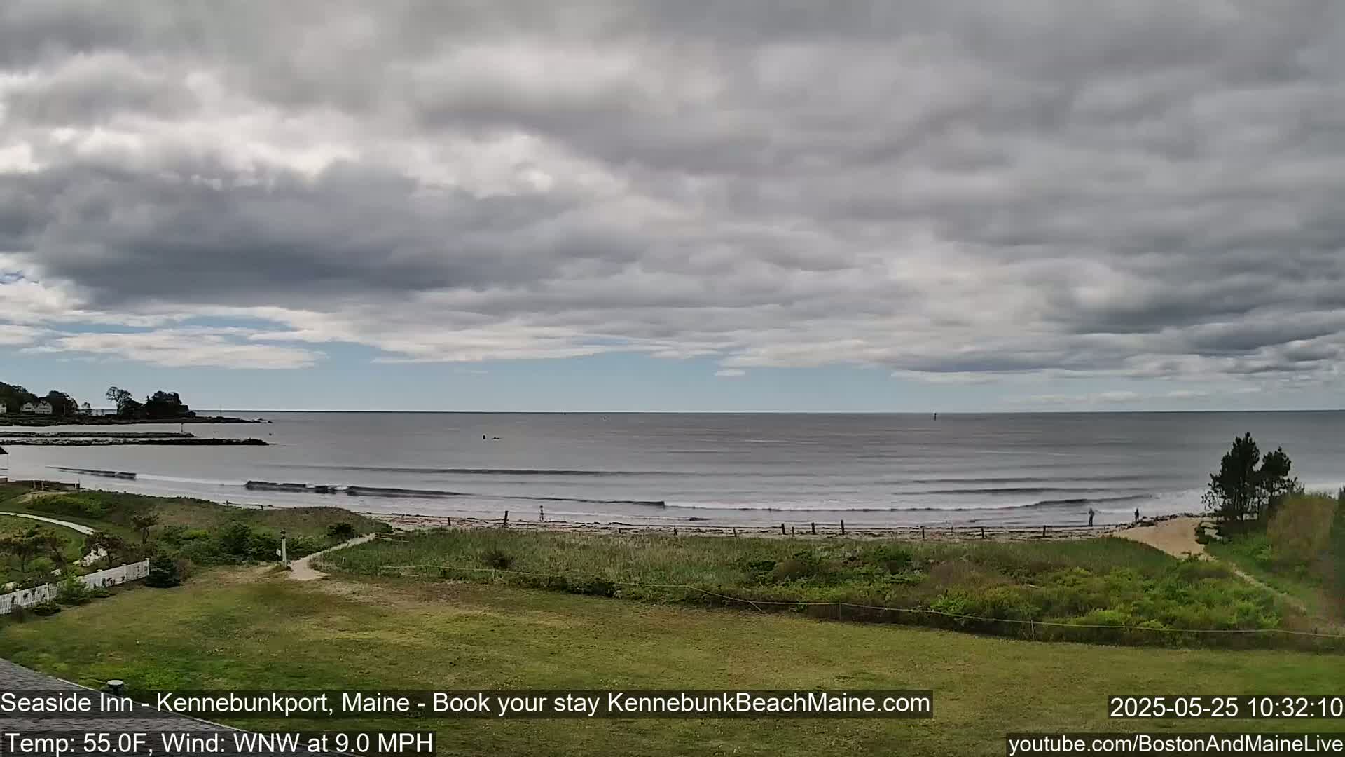 A mostly cloudy sky overlooks a calm ocean beach with small waves and green vegetation in the foreground.