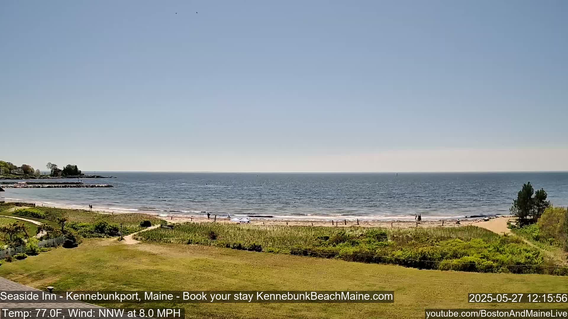A sandy beach with a few people on it stretches along a calm ocean under a clear blue sky.