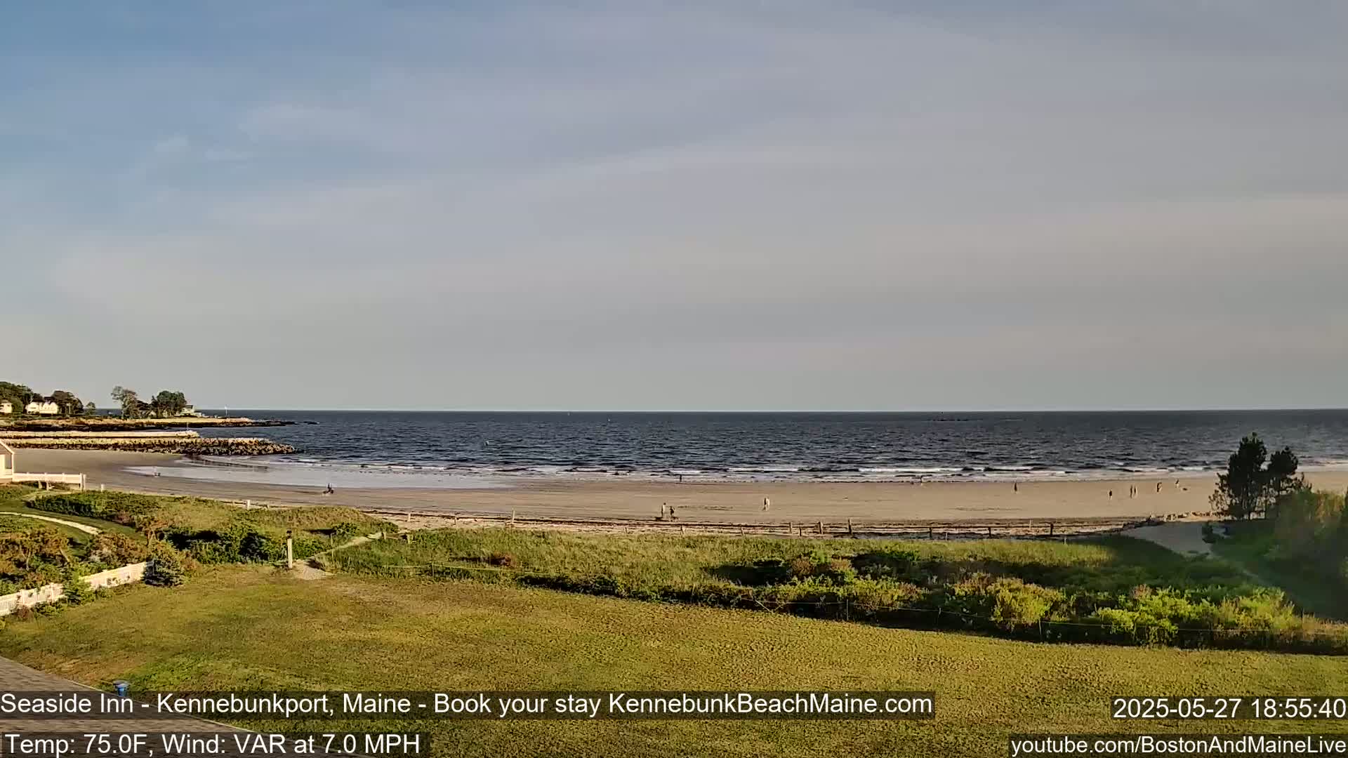 A sandy beach with a few people on it stretches along a calm ocean under a mostly clear sky.