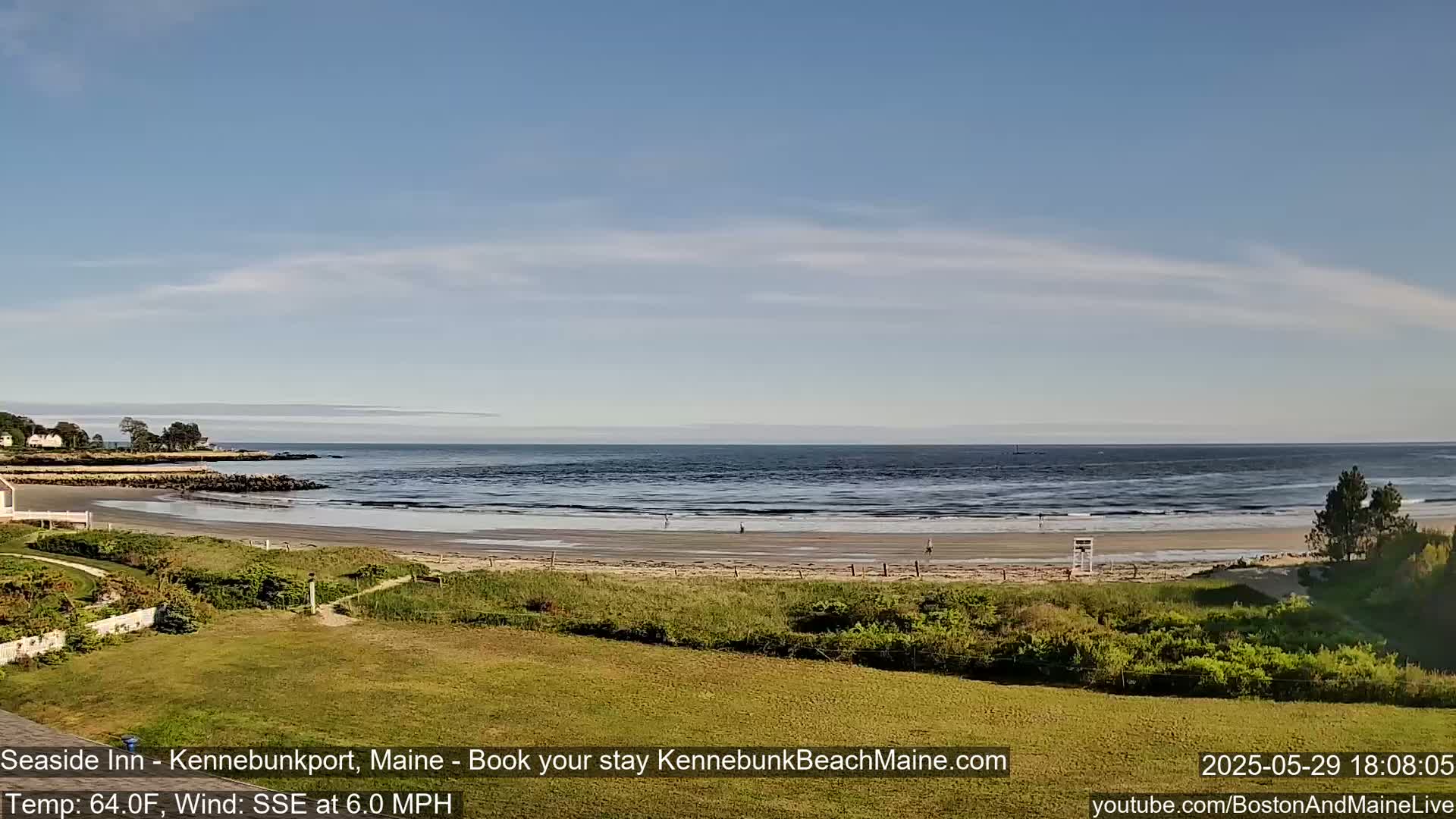 A sandy beach with a calm ocean under a mostly clear sky stretches out from a grassy area.