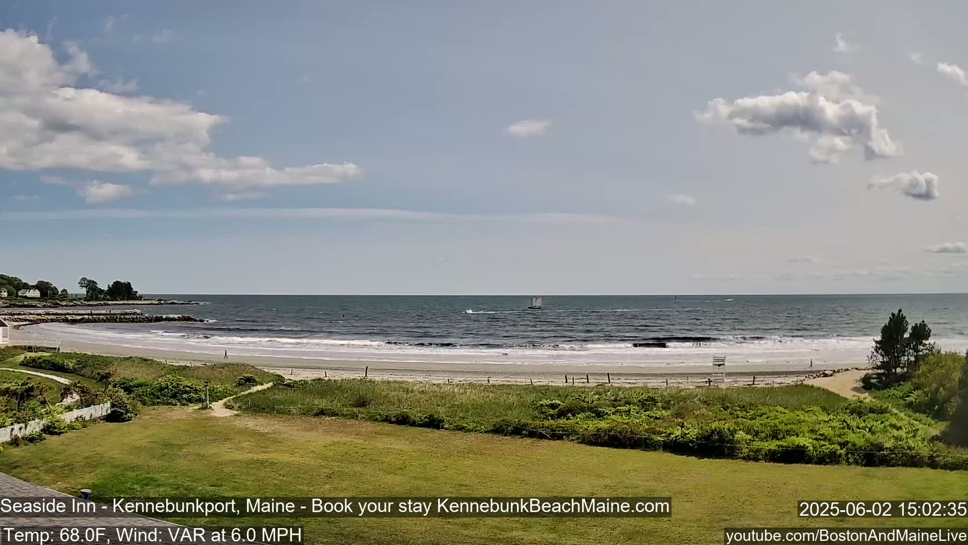 A sandy beach with small waves laps the shore under a partly cloudy sky; a sailboat is visible on the calm ocean in the distance.