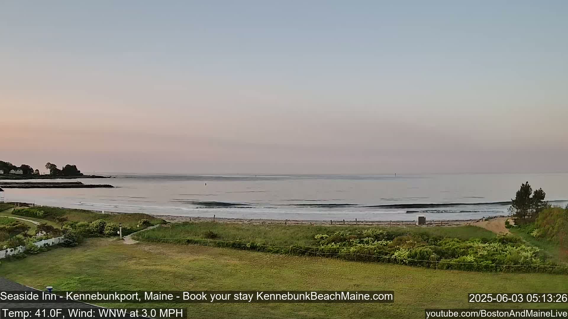 A calm ocean with small waves laps a sandy beach under a pale pink and gray sunrise.