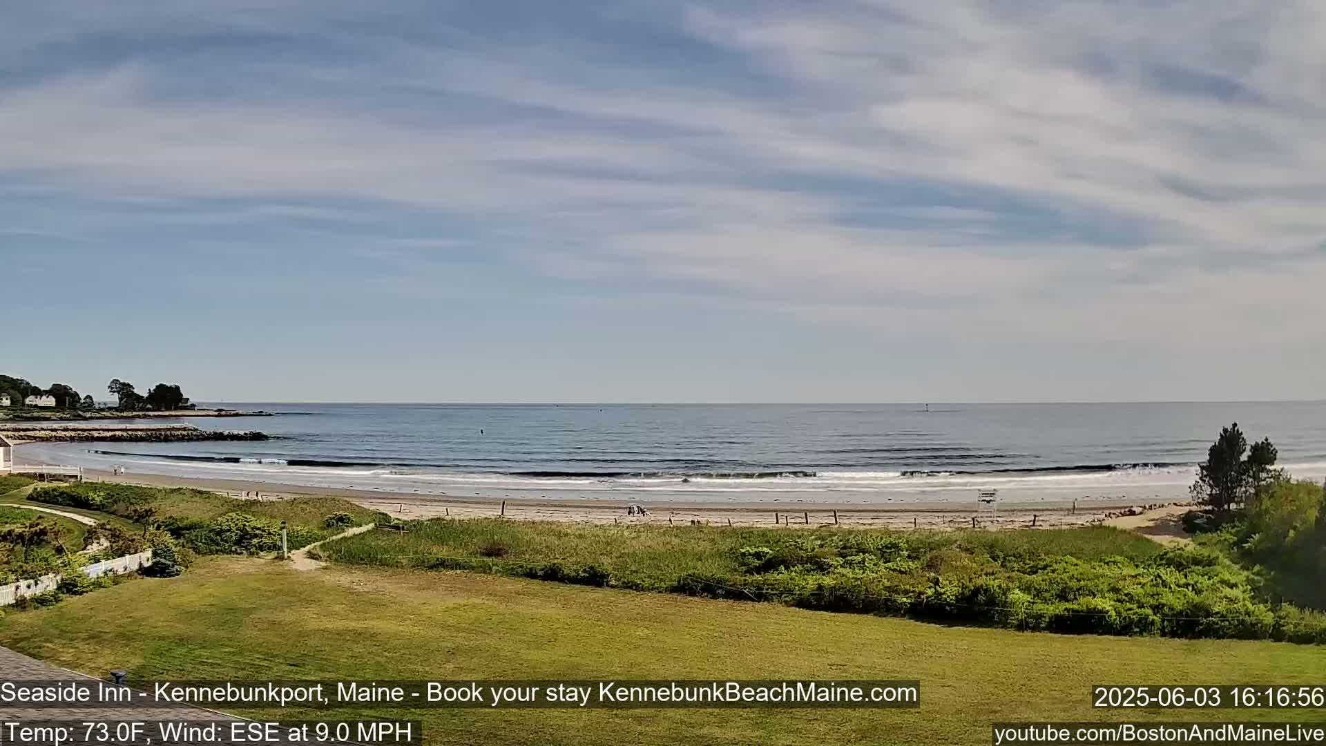A sandy beach with gentle waves and a few people stretches along a calm ocean under a partly cloudy sky.