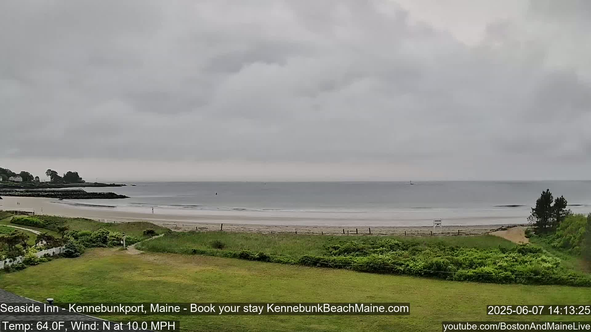 A long sandy beach with calm ocean waves is visible under an overcast sky, with green vegetation in the foreground.
