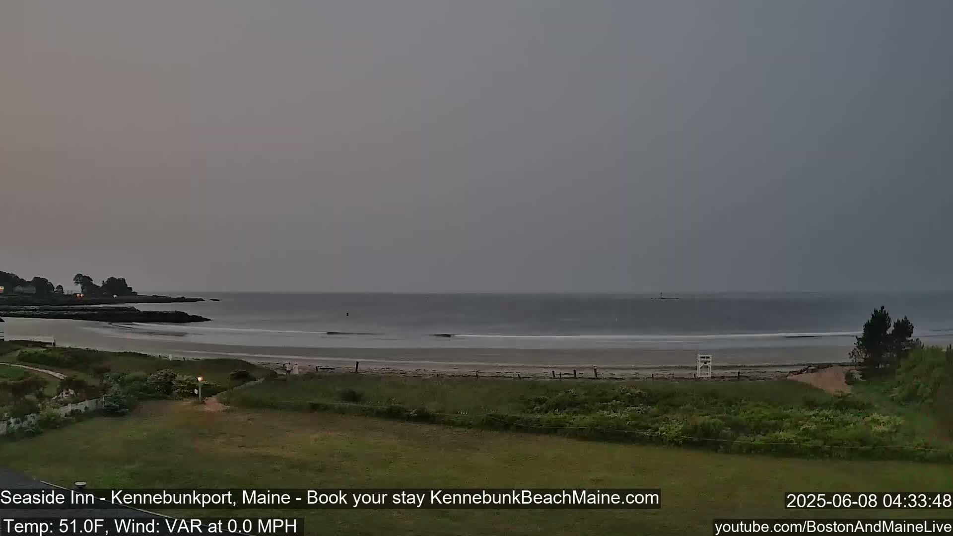 A calm, overcast ocean beach scene is shown with a lifeguard stand on the sand, and dark green vegetation in the foreground.