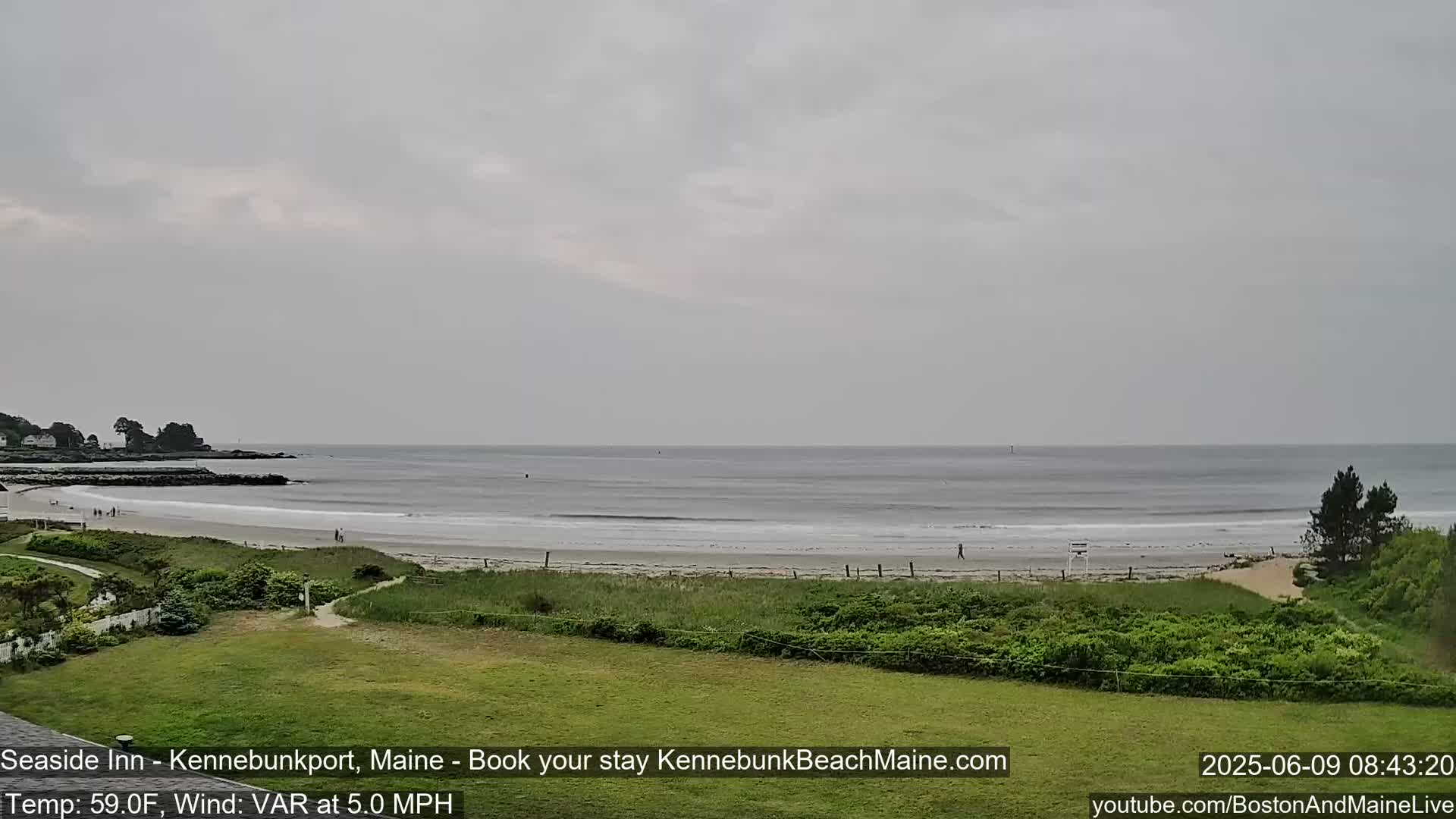 A sandy beach with a calm ocean under an overcast sky is viewed from a grassy area.