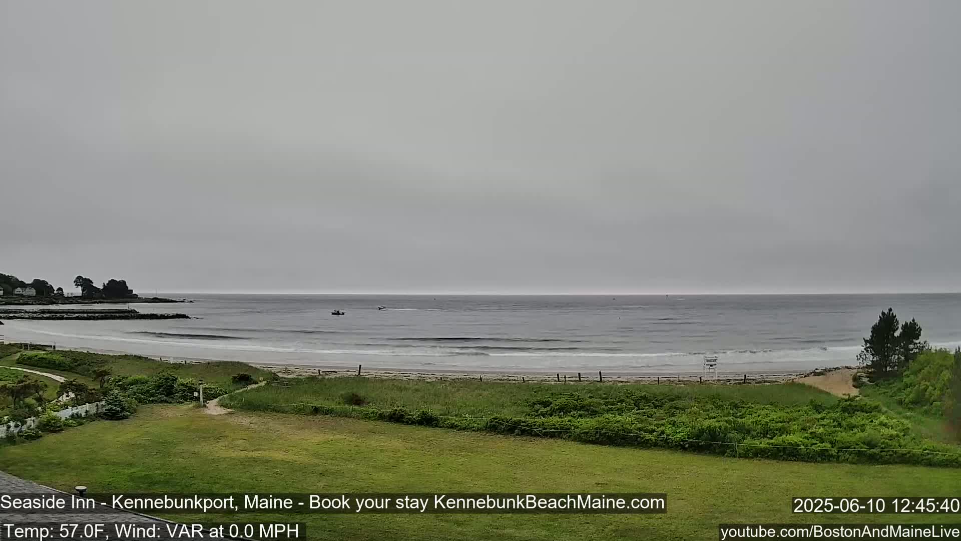 A calm, overcast day shows a sandy beach with small waves lapping the shore, a few small boats on the water, and green vegetation in the foreground.