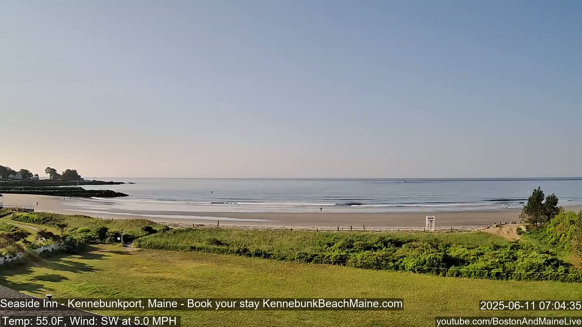 A sandy beach with calm waves and a lifeguard stand is visible from a grassy area overlooking the ocean on a clear, sunny morning with a southwest wind.