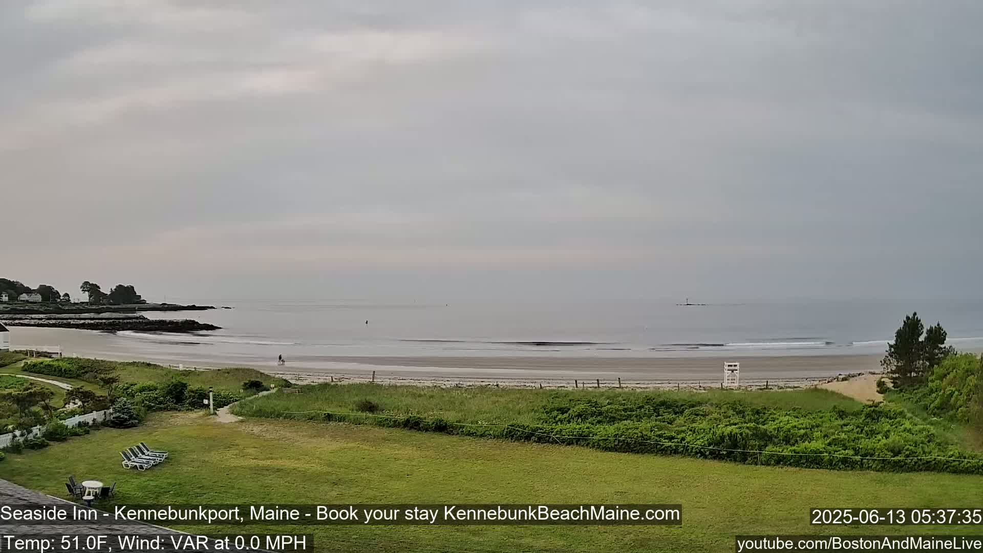 A calm, overcast day reveals a sandy beach with a lifeguard stand, extending to a tranquil ocean with a distant rocky outcrop.