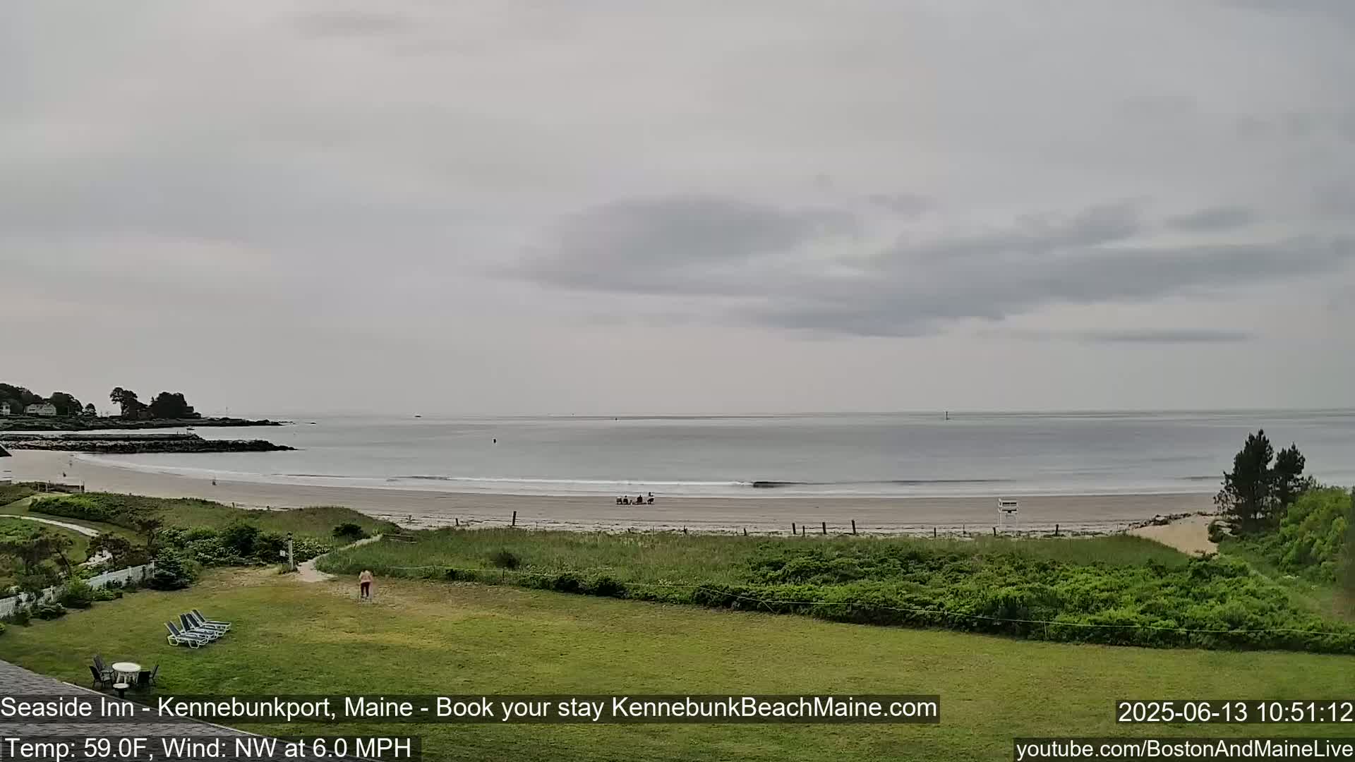 A calm, overcast day reveals a sandy beach and calm ocean, viewed from a grassy area with lounge chairs.