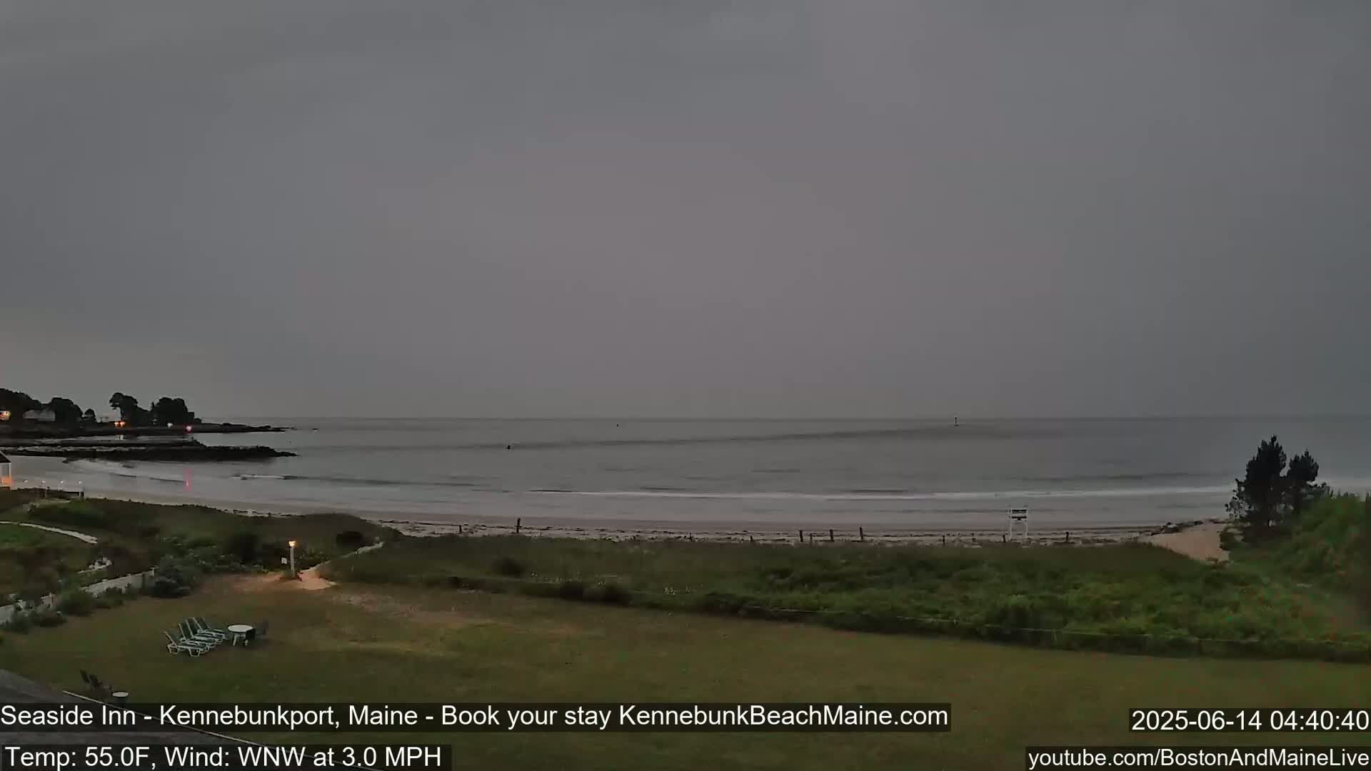 A calm ocean beach is seen under an overcast sky, with a grassy area and some trees in the foreground.