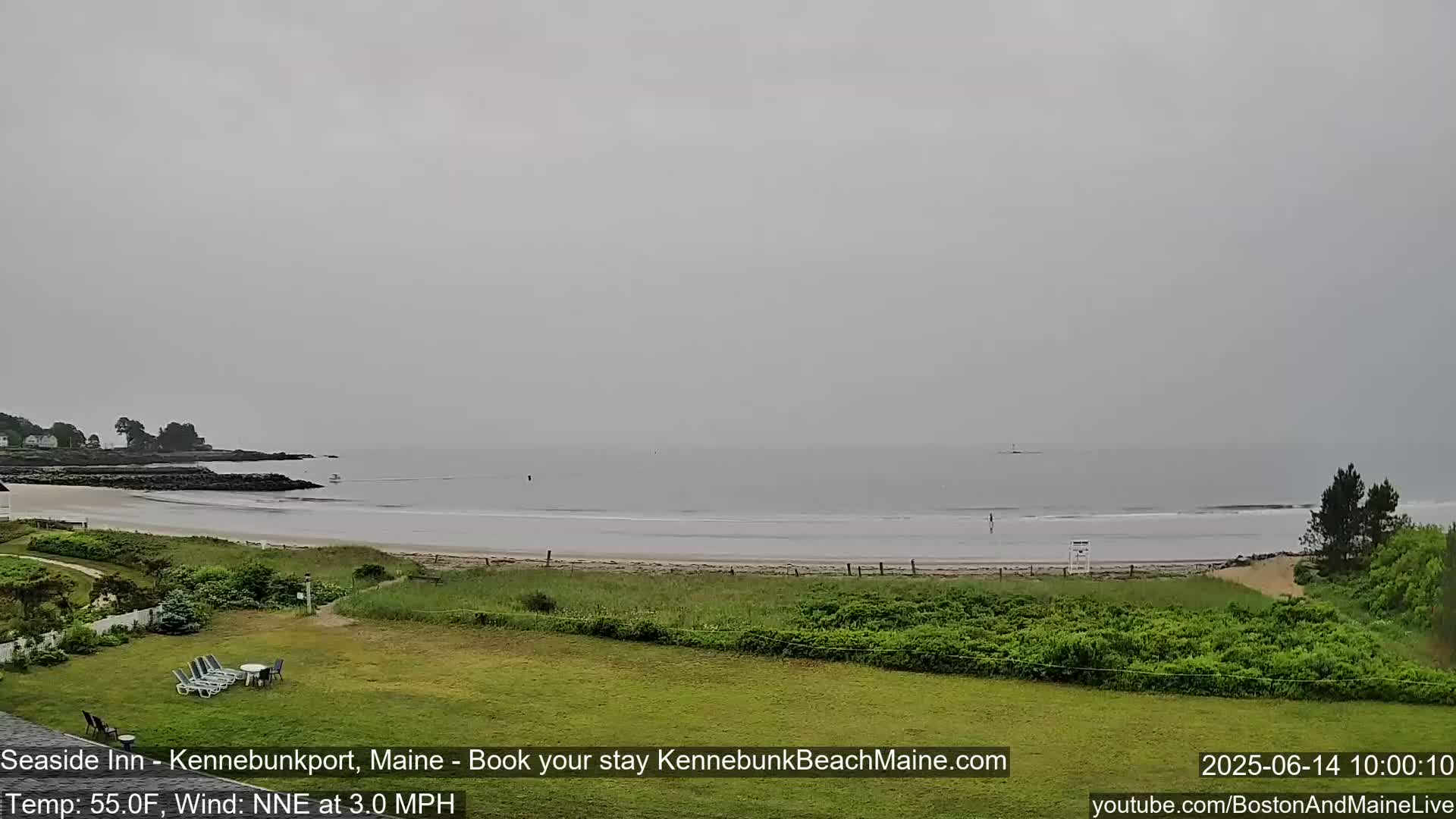 A hazy, overcast day reveals a beach scene with calm waves, a sandy shore, grassy dunes, and a distant rocky coastline.