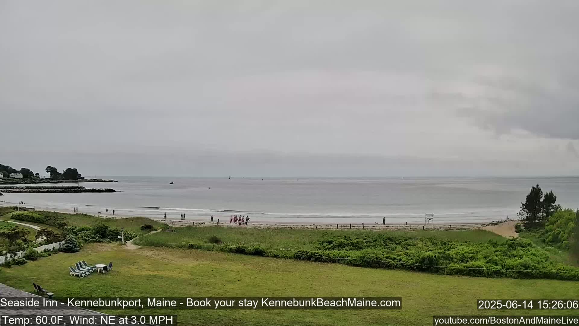 A hazy, overcast day reveals a beach scene with calm waves, a sandy shore, grassy dunes, and a distant rocky coastline.