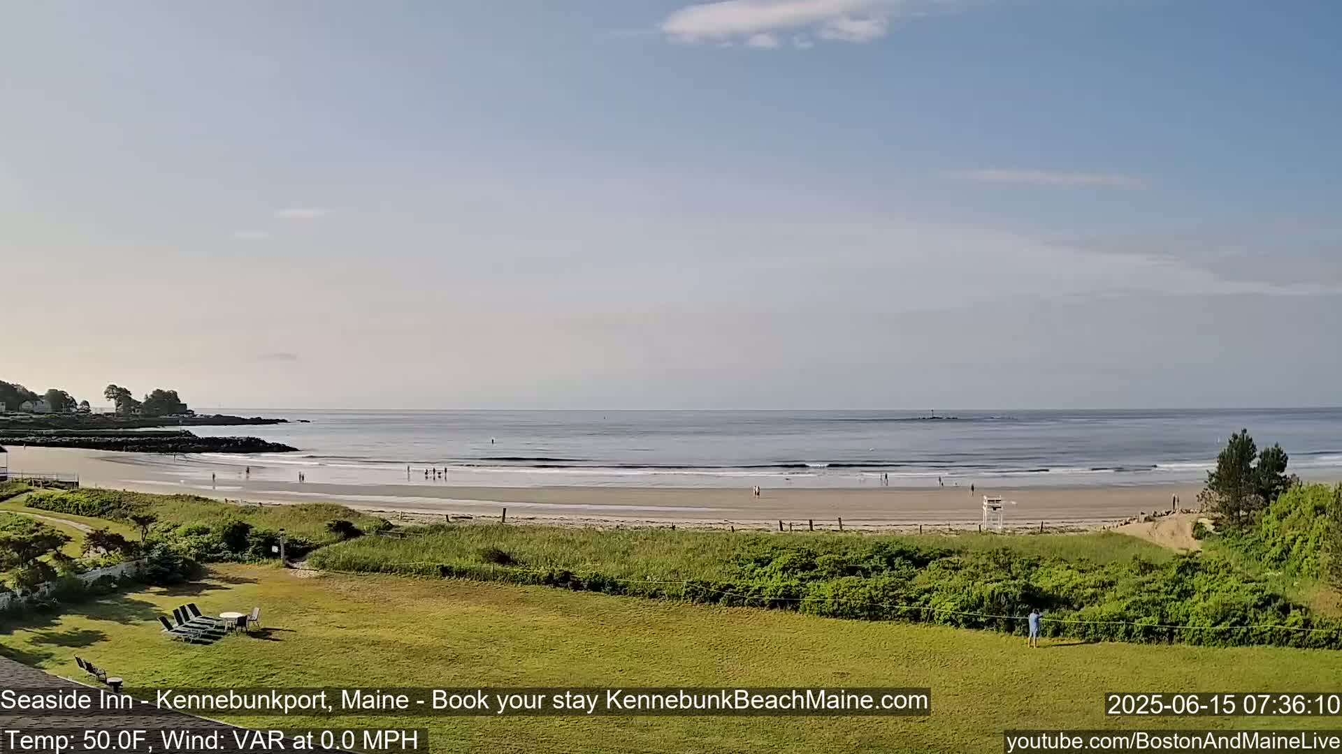 A calm ocean with small waves laps a sandy beach where a few people are scattered, all under a mostly clear, light blue sky.