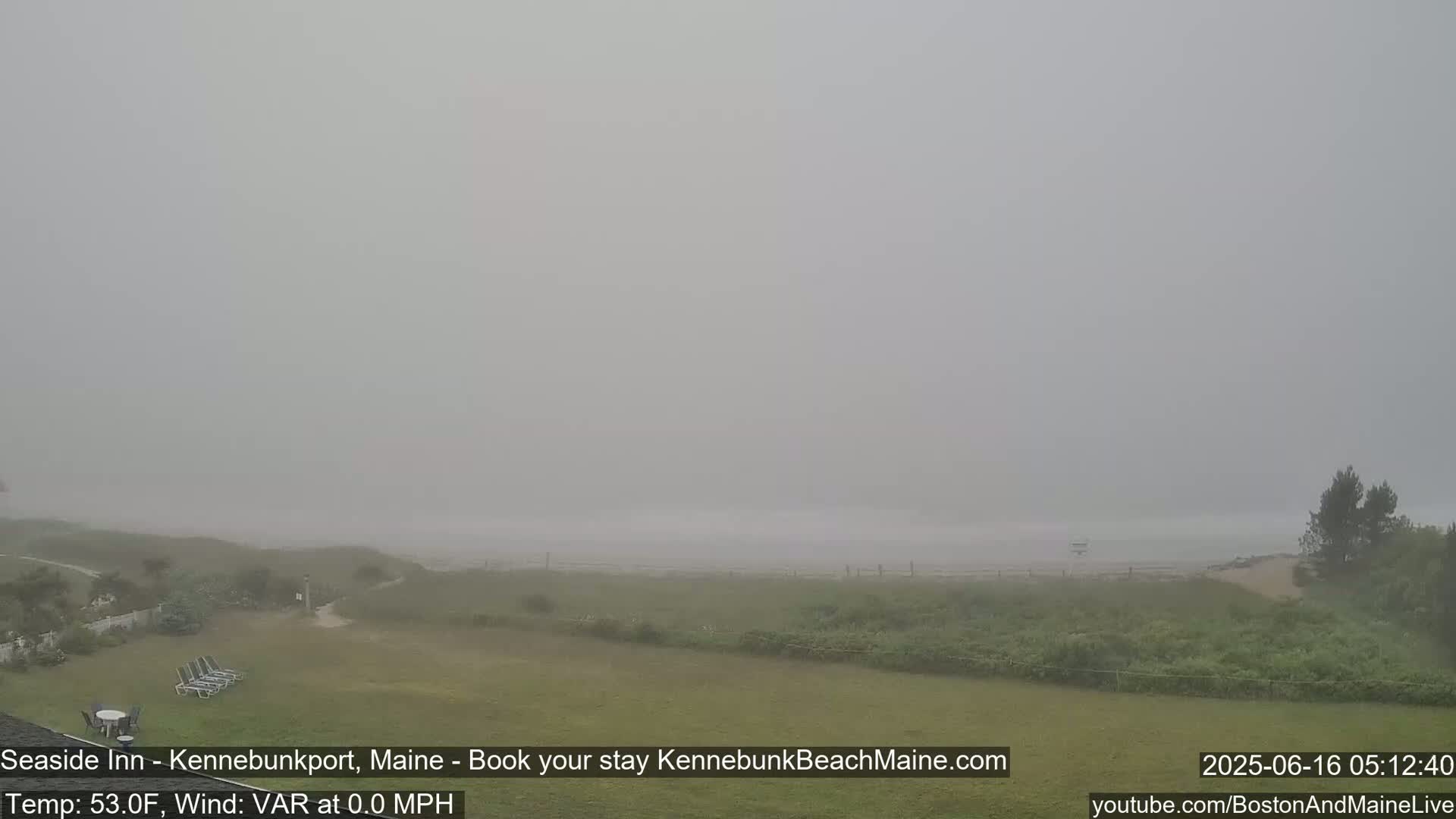 A foggy seaside scene shows a grassy lawn with beach chairs, leading to a hazy beach and ocean under a gray sky.