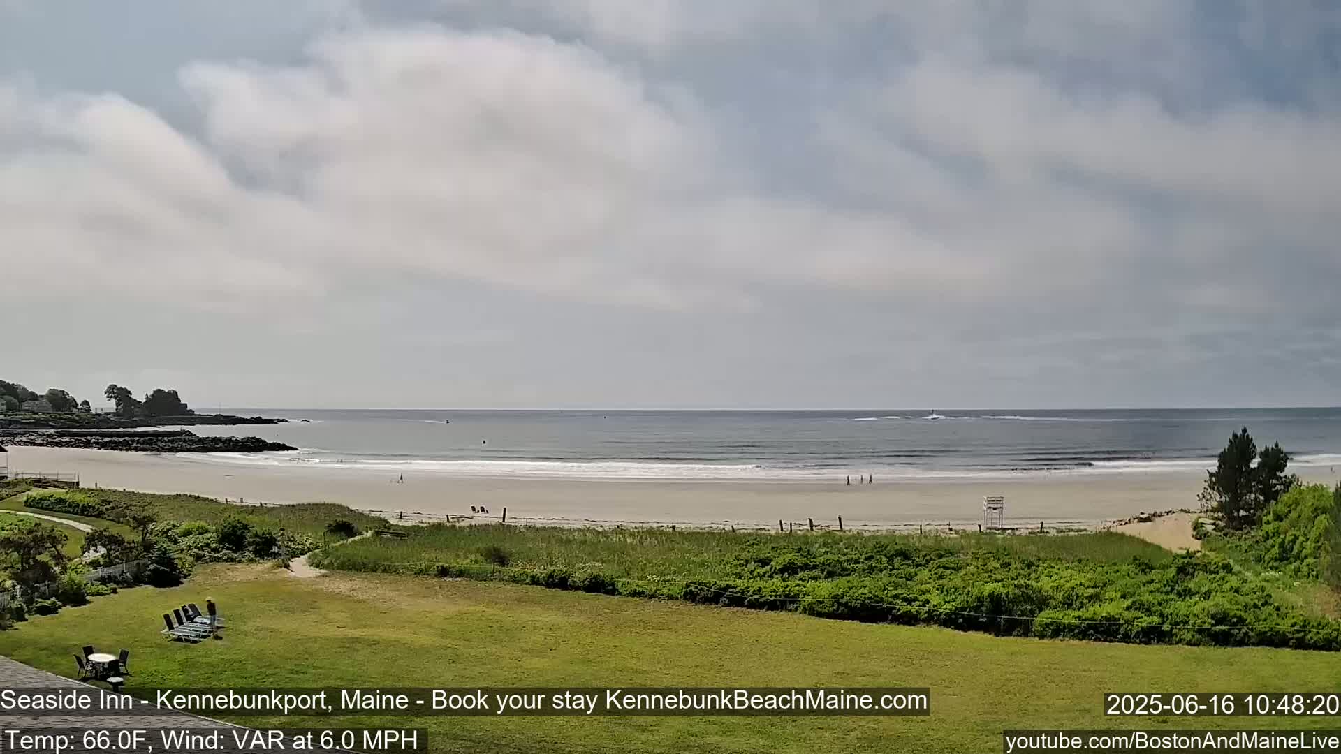 A sandy beach with a calm ocean stretches out under a mostly cloudy sky, viewed from a grassy area with some shrubbery.