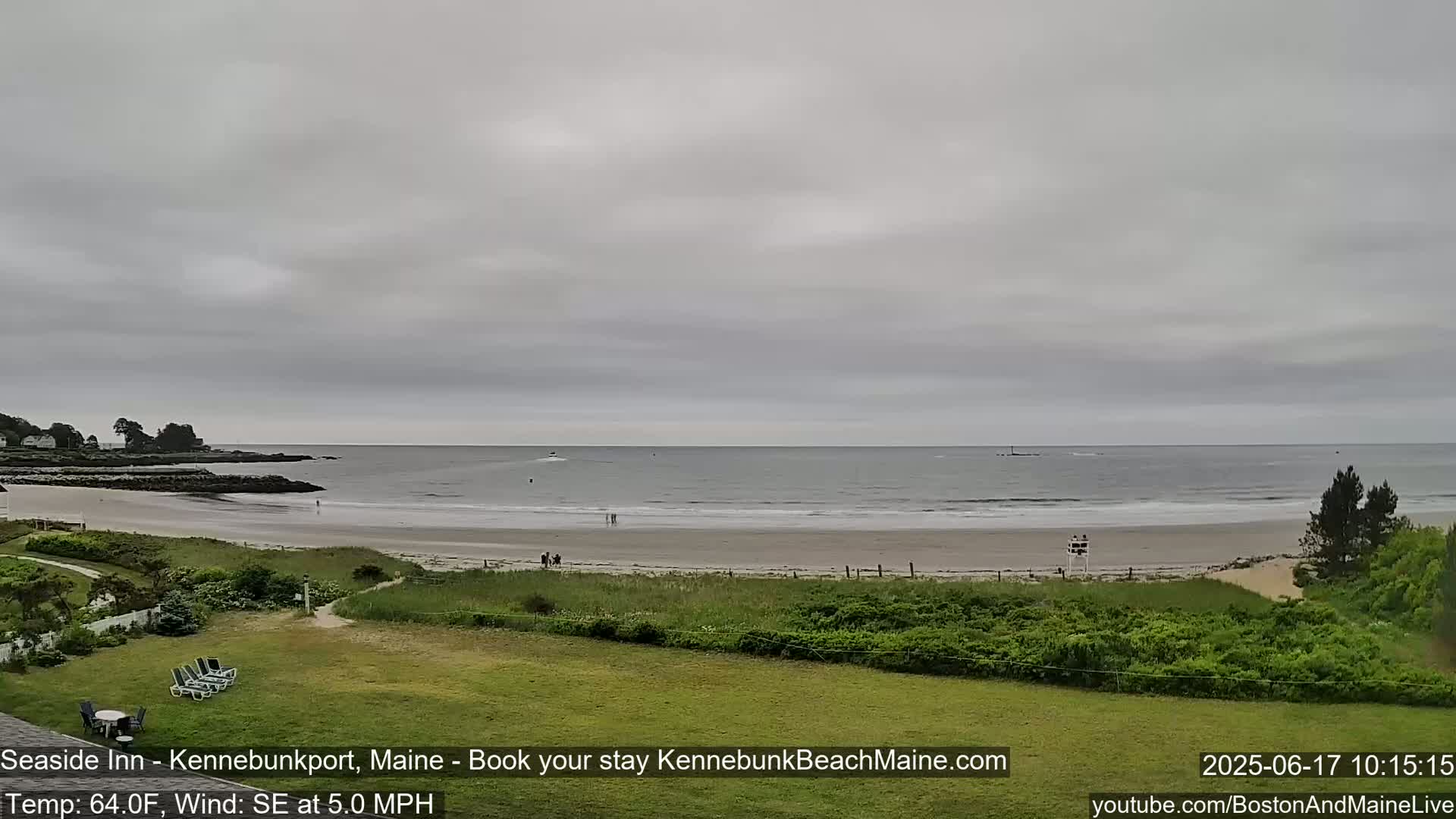 A view of a sandy beach with calm waves under an overcast sky, showing a few people on the shore and some vegetation in the foreground.