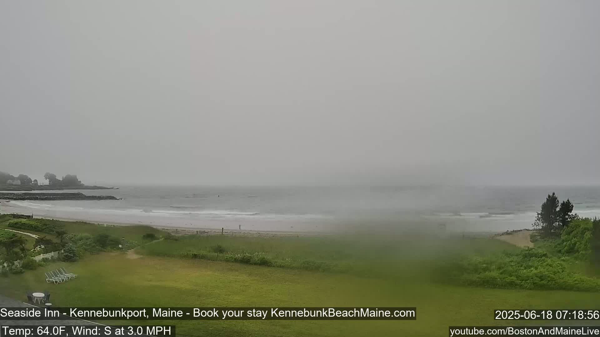 A hazy, overcast view shows a beach with a calm ocean, some vegetation, and a grassy area in the foreground.